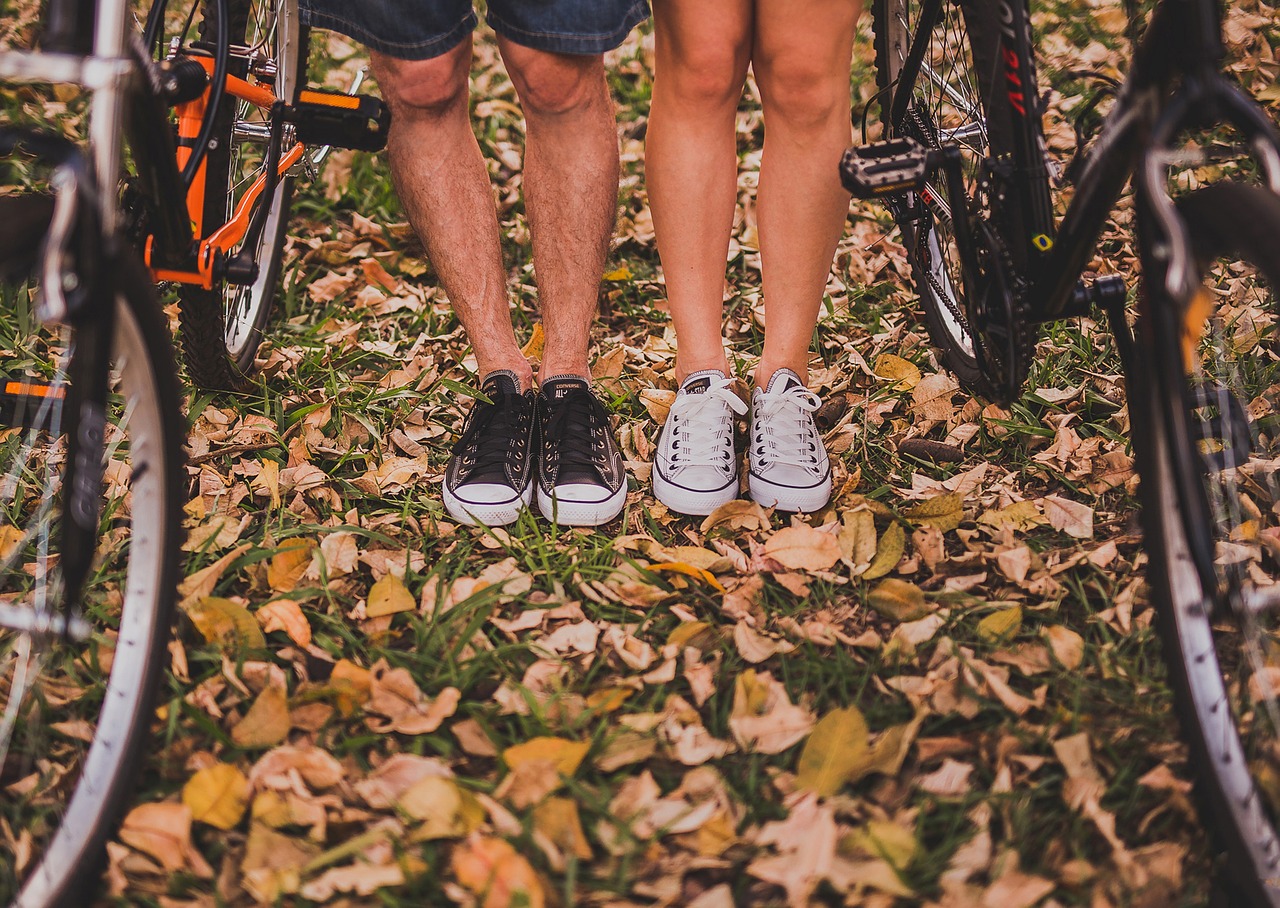homme et femme dont on ne voit que les jambes à partir du genou portant des converses noires et blanches à côté de vélos sur sol plein de feuilles mortes