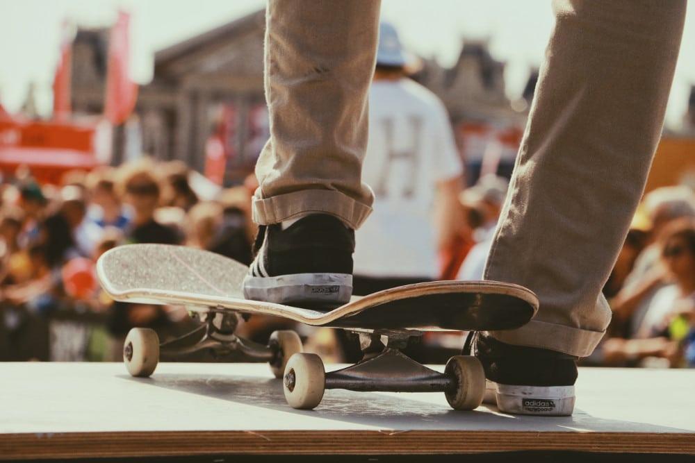 vue sur les pieds d'un homme portant un chino beige et des baskets foncées un pied posé sur un skate